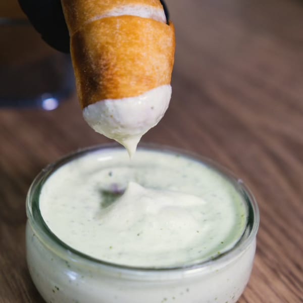 Tequeño being dipped into a creamy green sauce, with a drip falling back into the bowl.