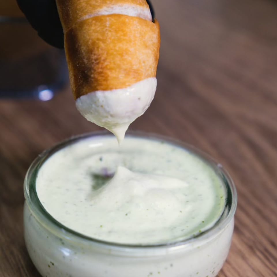 Tequeño being dipped into a creamy green sauce, with a drip falling back into the bowl.