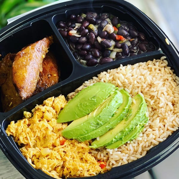Venezuelan meal with plantains, black beans, brown rice, scrambled eggs, and sliced avocado in a to-go container.