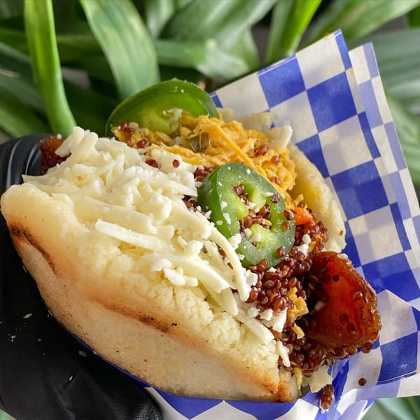 A Venezuelan arepa filled with shredded cheese, quinoa, plantains, and jalapeños, held in checkered paper.