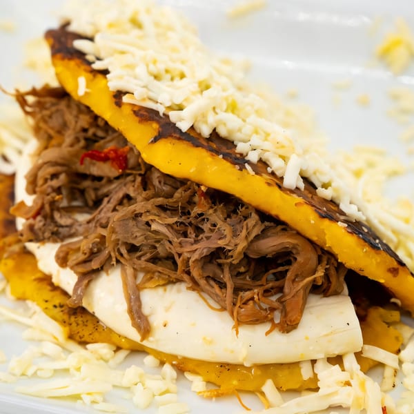 Close-up of a Venezuelan arepa with shredded beef, white cheese, and plantain on a white plate.