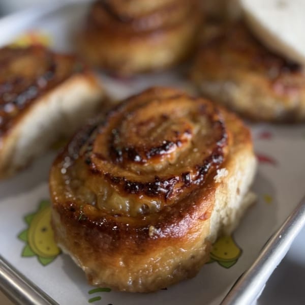 Close-up of a golden-brown, glazed cinnamon roll on a white tray with cartoon avocado illustrations.